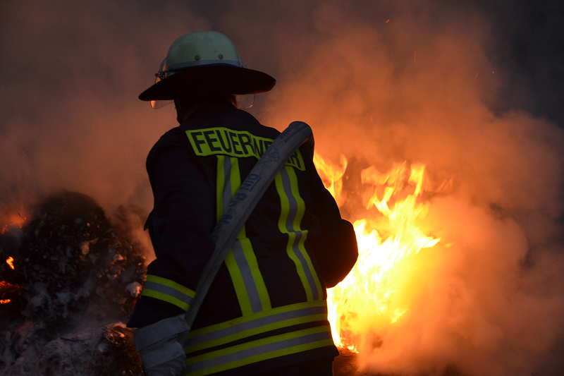Ein Feuerwehrmann in Schutzkleidung und Helm bekämpft einen offenen Brand mit einem Löschschlauch. Dichte Rauchschwaden und helle Flammen erhellen die Szene.