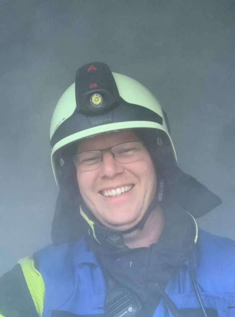 Ein lächelnder Feuerwehrmann mit Helm und Brille blickt in die Kamera. Leichter Rauch umgibt die Szene, was auf eine Einsatzübung oder einen realen Einsatz hindeutet.